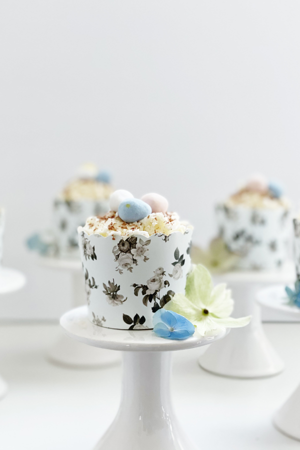 Beautifully styled Easter egg cupcakes sit on crisp white pedestals, each wrapped in a scalloped black floral liner with intricate botanical details. The velvety white buttercream is dusted with rich chocolate curls and crowned with pastel mini eggs, making this a showstopping Easter-themed dessert for any celebration.