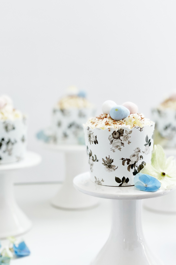 A delicate spring cupcake idea rests on a classic white pedestal, its soft white frosting swirled to perfection and adorned with chocolate shavings. Wrapped in a scalloped black floral liner with intricate botanical details, this cupcake is a picture-perfect Easter dessert idea for spring celebrations.