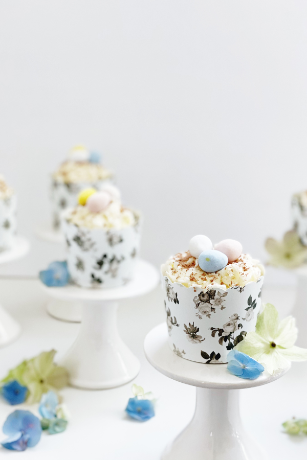 Easter cupcakes for kids and adults alike are displayed on a white pedestal. They are wrapped in a scalloped black floral cupcake liner with hints of blue and green florals scattered around. The easy Easter cupcakes are topped with a swirl of white buttercream, delicate chocolate curls, and pastel mini eggs, this Easter cupcake idea is both timeless and festive.