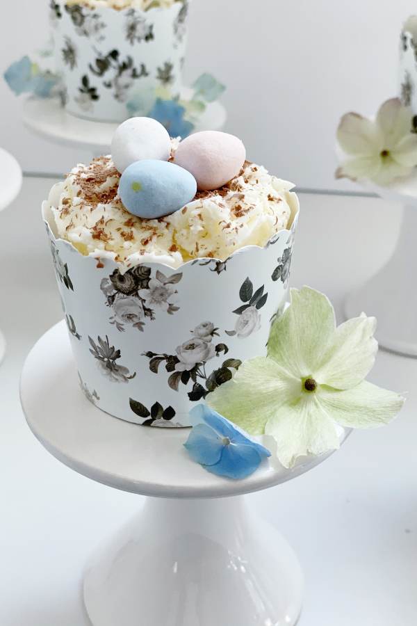 A charming Easter dessert displayed on an elegant white pedestal, featuring a scalloped black floral cupcake wrapper. The cupcake is crowned with fluffy white frosting, fine chocolate shavings, and soft-hued mini eggs, making this an easy Easter cupcake that blends timeless beauty with seasonal delight.