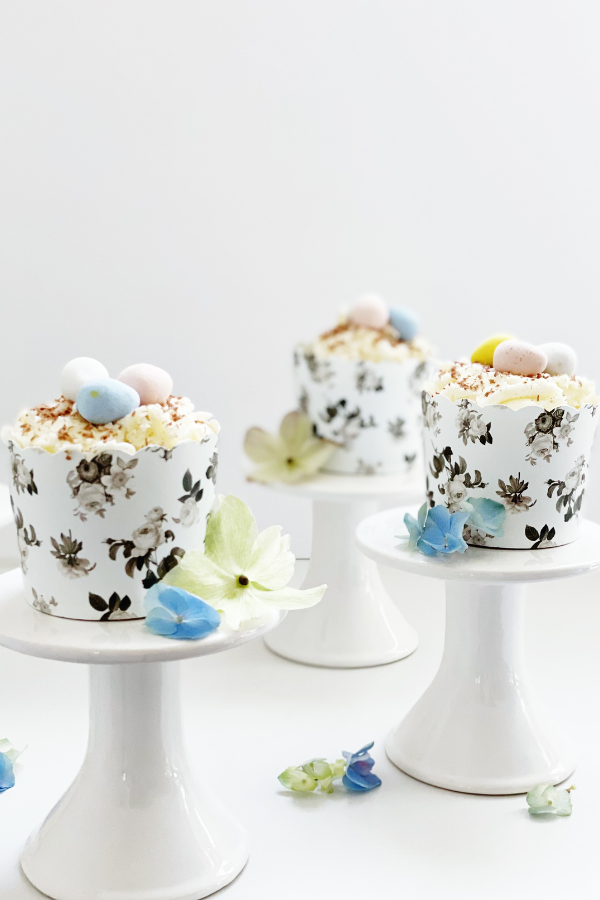 A charming Easter dessert idea featuring a chocolate cupcake with fluffed white frosting, fine chocolate shavings, and soft pastel mini eggs. Nestled in a sophisticated black floral wrapper and placed on a white pedestal. These cute Easter desserts blend rustic elegance with seasonal charm.