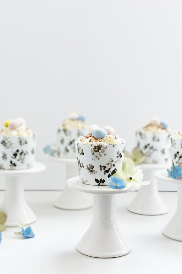 A sophisticated Easter cupcake idea presented on a white pedestal, wrapped in an intricate black floral wrapper. The cupcake’s smooth white frosting is accented with delicate chocolate curls and speckled pastel mini eggs, creating an effortlessly elegant Easter-themed dessert for spring gatherings.